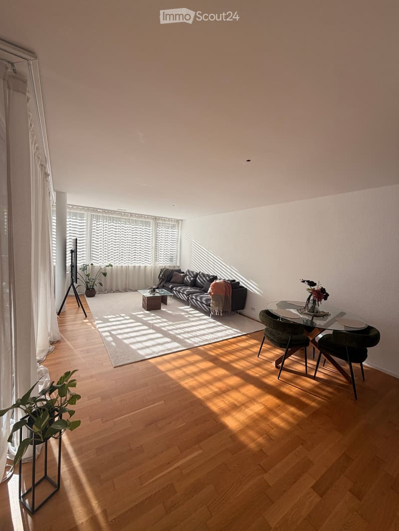 Bright living room with wooden flooring, large windows with white curtains, a black sectional sofa, a glass coffee table, a dining table with chairs, and plants.