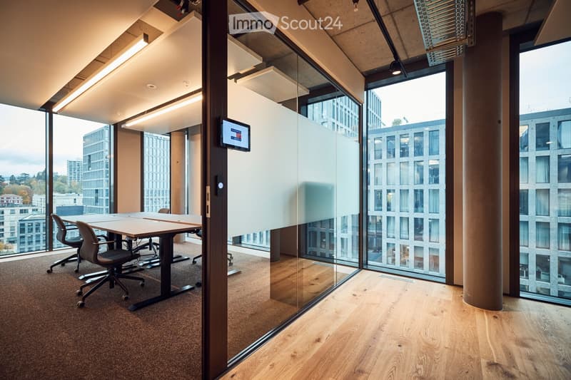 Modern office space with large glass windows, a wooden table, and chairs, featuring a sleek design and natural light.