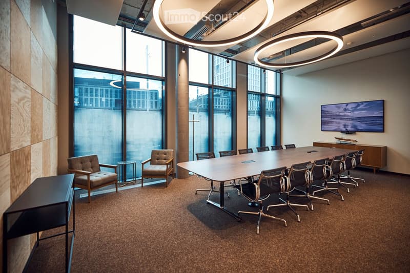 Modern conference room with large windows, a long conference table surrounded by office chairs, side chairs, a small round table, and a screen mounted on the wall.