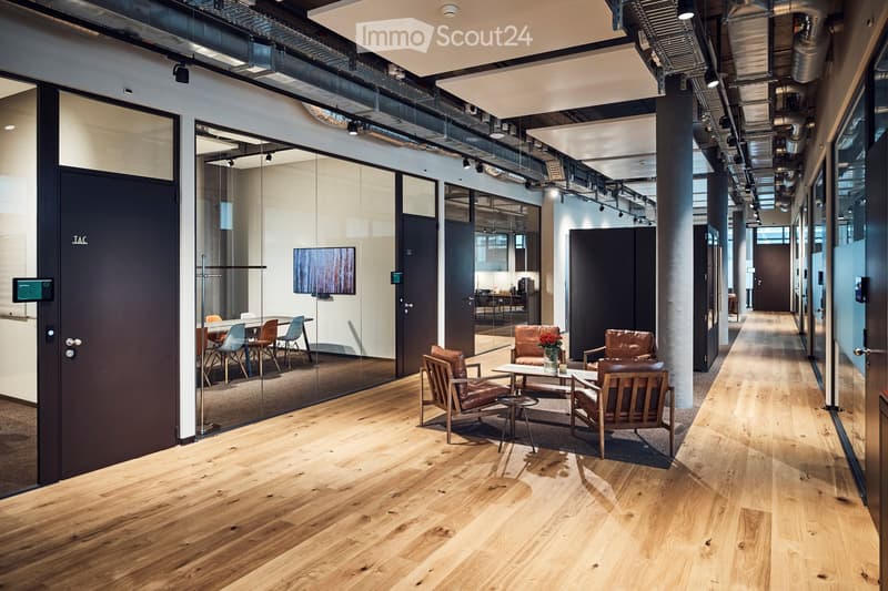 Modern office space with wooden flooring, glass partitions, leather chairs, small tables, and a screen in the background.