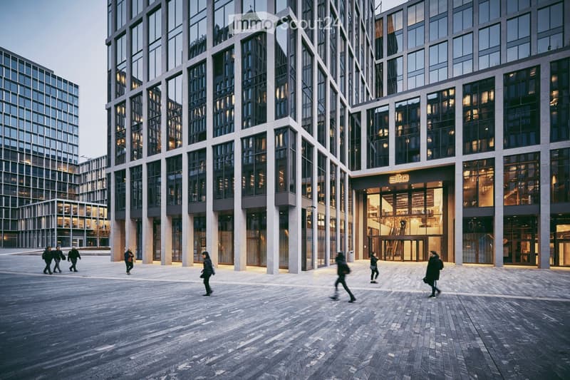 Modern multi-story building with large glass windows and a central entrance, surrounded by a paved plaza with people walking.