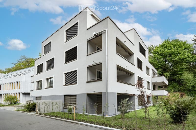 Modern multi-story building with white facade, large windows, and balconies. Features a garage entrance and landscaped surroundings with greenery.