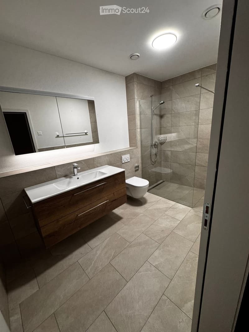Modern bathroom with beige tiles, a wooden vanity with a sink, a toilet, and a glass-enclosed shower area.