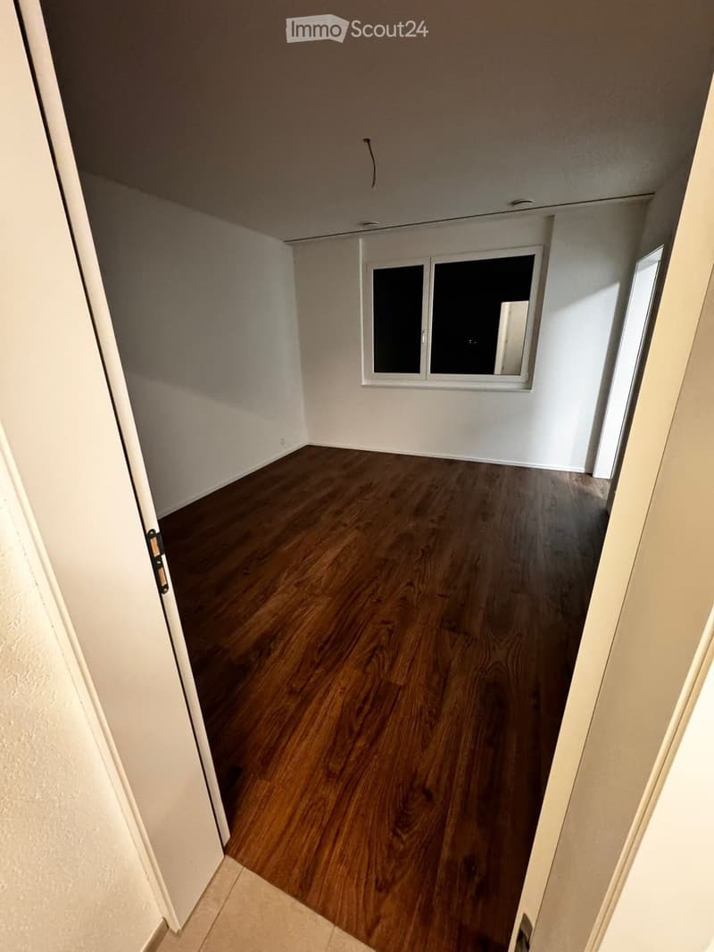Empty room with wooden flooring, white walls, and a window with two panes. The room is unfurnished and has a door visible on the left side.