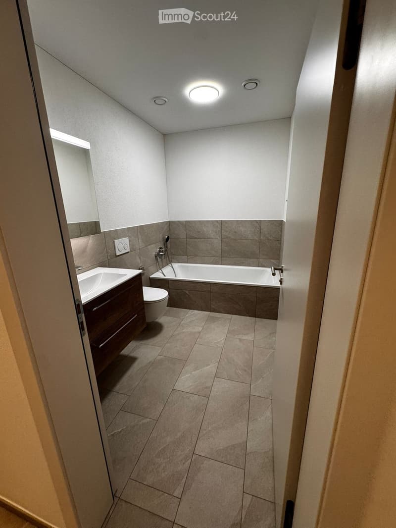 Modern bathroom with a bathtub, toilet, sink, and wooden cabinet. Features tiled walls and floor, recessed lighting, and a built-in shelf.