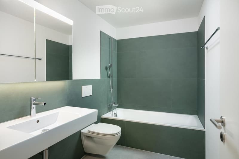 Modern bathroom with a white sink, chrome faucet, toilet, bathtub, and showerhead. Green tiles on the walls, white cabinetry, and a large mirror above the sink.