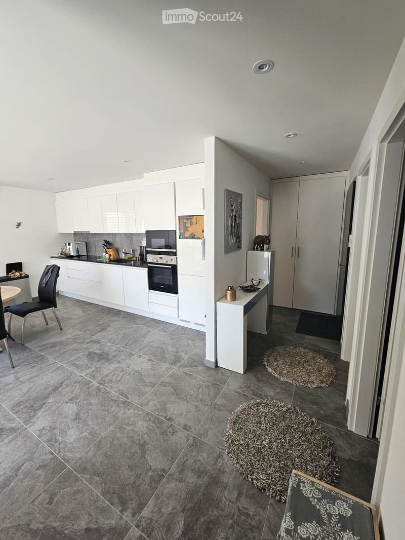 Modern kitchen with white cabinets, stainless steel appliances, and a dining area with a round table and chairs. The floor is tiled in grey marble, and there are two small round rugs in the seating area.