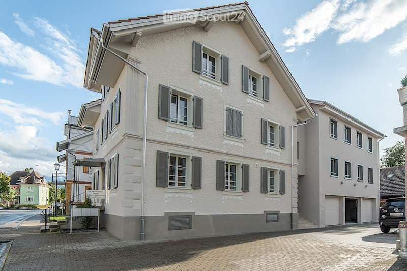 A two-story residential house with white siding, dark shutters, and multiple windows. The house features a sloped roof and a small entrance area with a signboard.