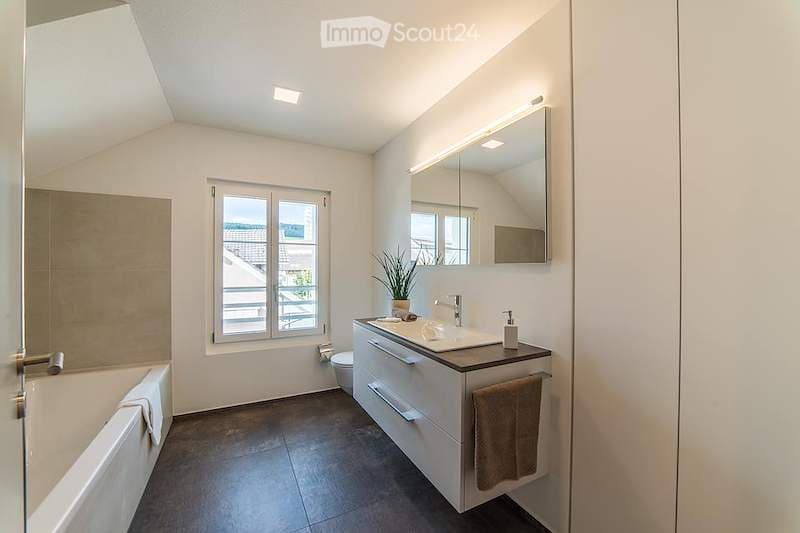 Modern bathroom with a bathtub, sink, and toilet. Features a large mirror with built-in lighting, a window with a view, and a small potted plant on the sink counter.