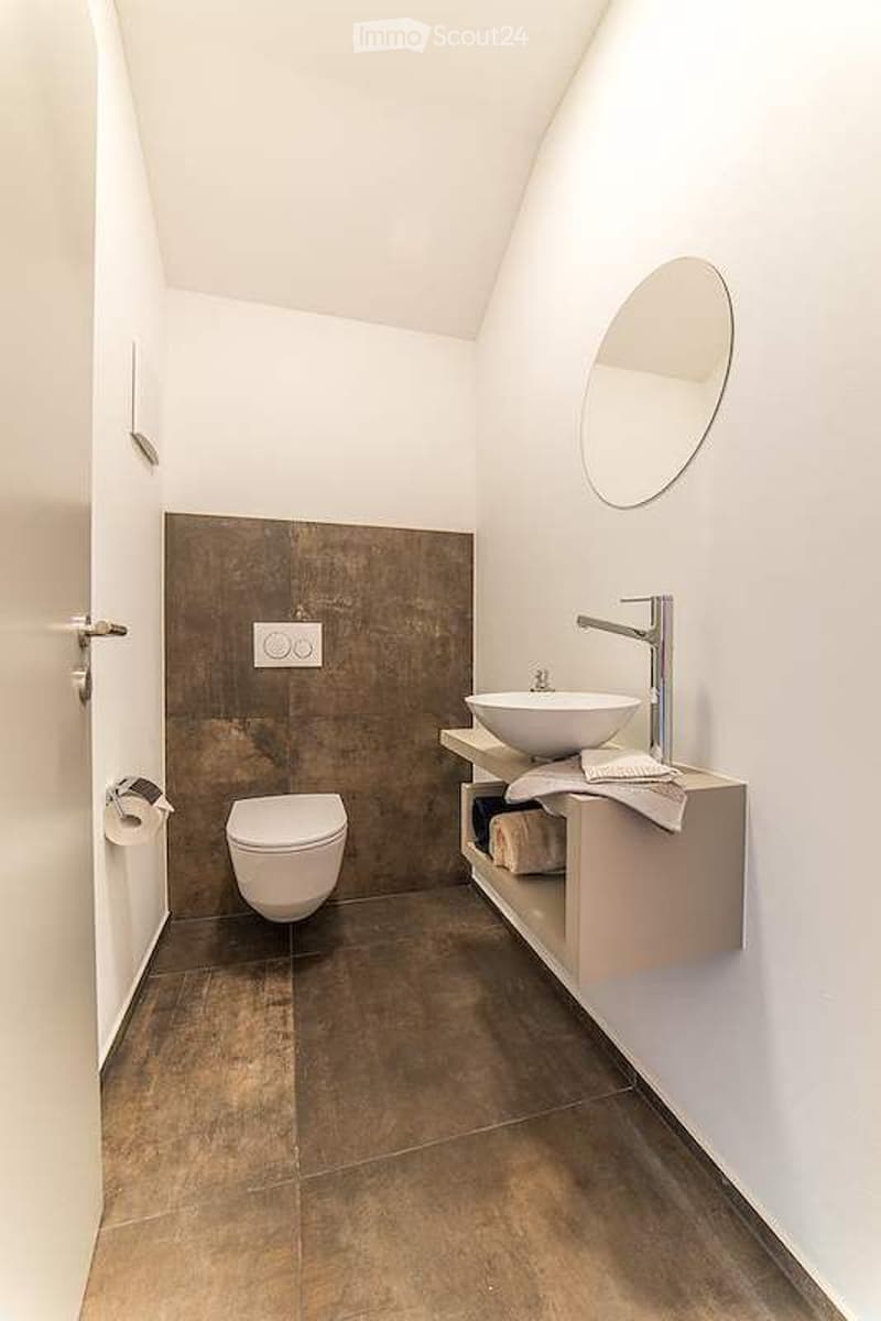 Modern bathroom with a dark brown accent wall, white tiles, a floating vanity with a sink, a toilet, and a round mirror above the sink.