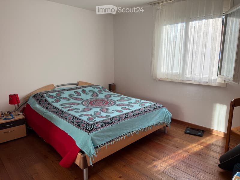 Cozy bedroom with wooden flooring, a bed with a colorful patterned bedspread, a nightstand with a lamp, and a chair near the window.
