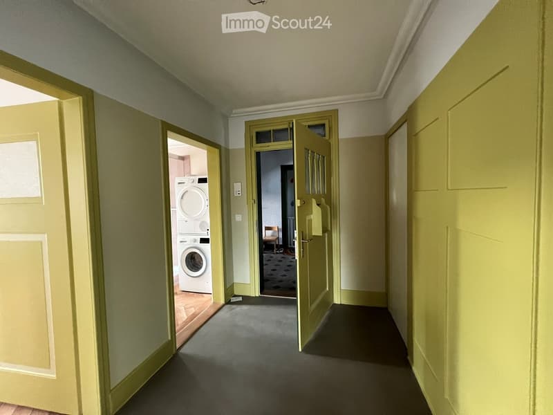 A narrow hallway with light green walls and carpeted floor, featuring two open doors leading to adjacent rooms, one showing a washing machine and dryer unit.