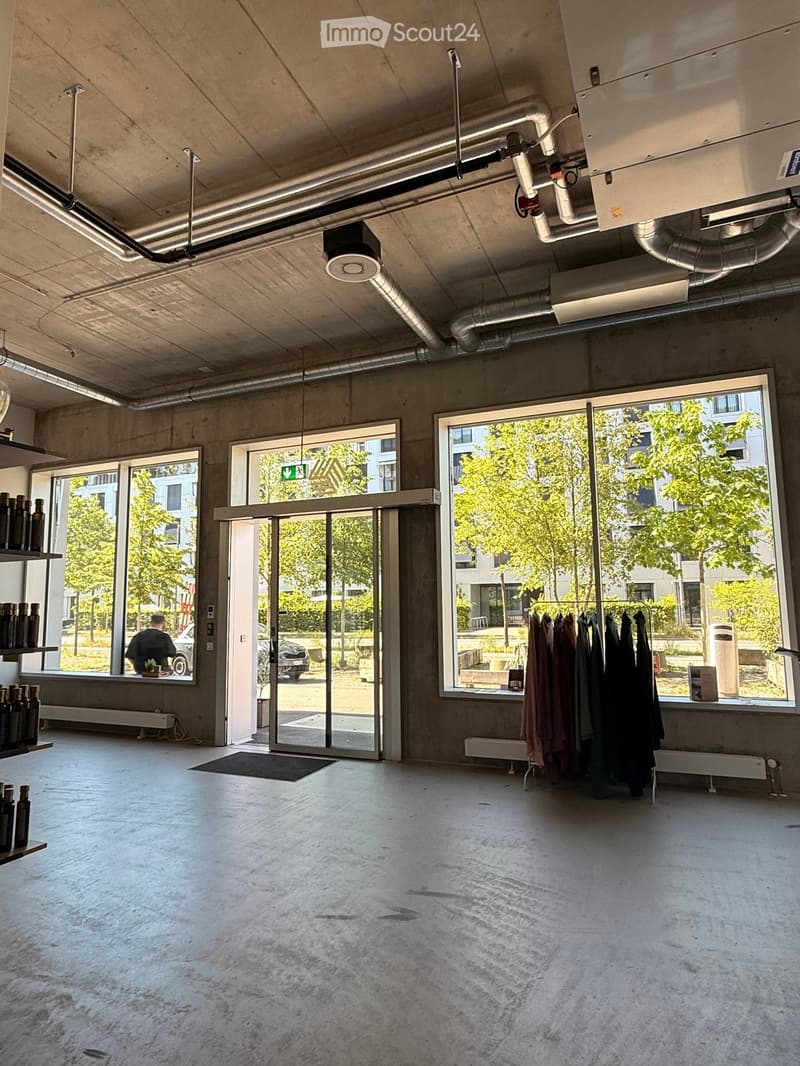 An empty, modern room with concrete floors and walls, large windows, and exposed pipes and lighting fixtures on the ceiling. Shelves with bottles and a clothing rack are visible, but the room itself is unfurnished.