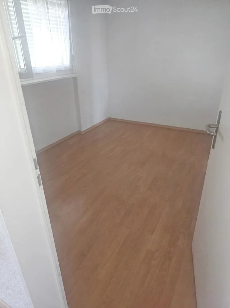 Empty room with wooden flooring and white walls, featuring a window with blinds and a door.