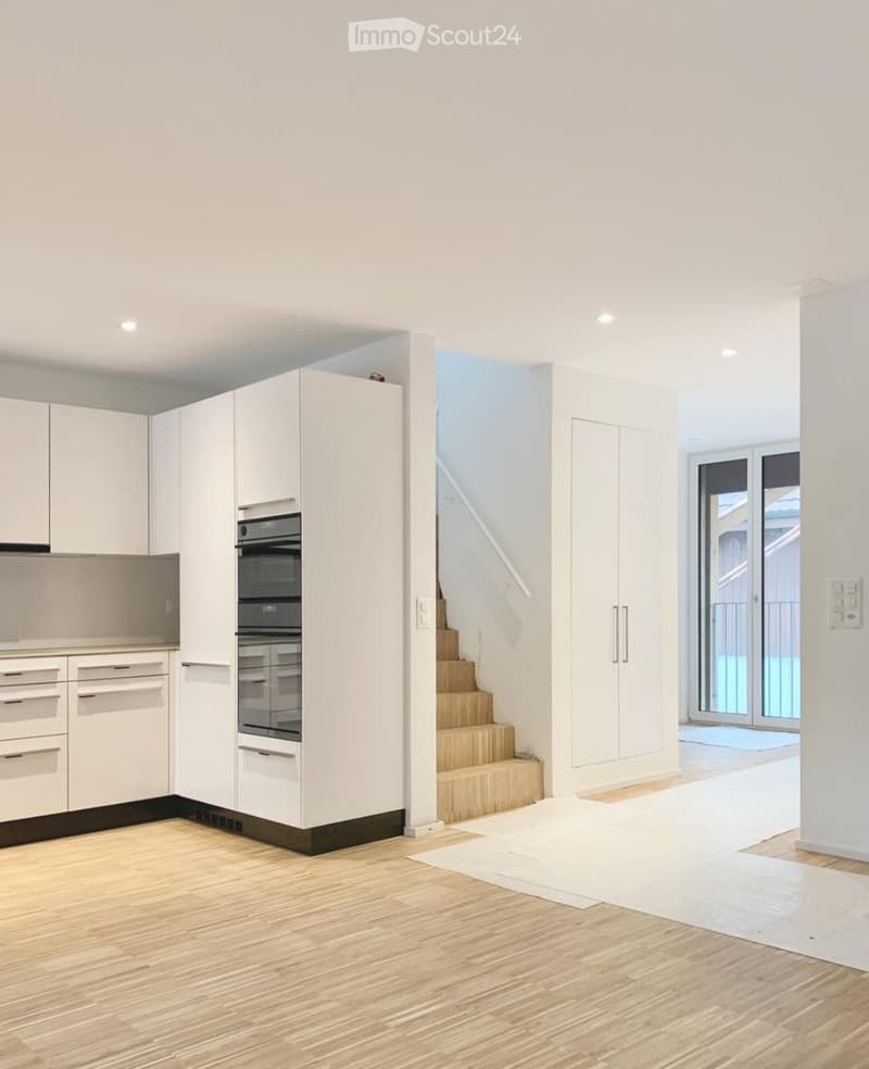 Open-plan space with modern white kitchen cabinets, built-in oven, and a staircase leading to an upper level. The floor is light wood, and there is a large sliding glass door leading to a balcony or outdoor area.