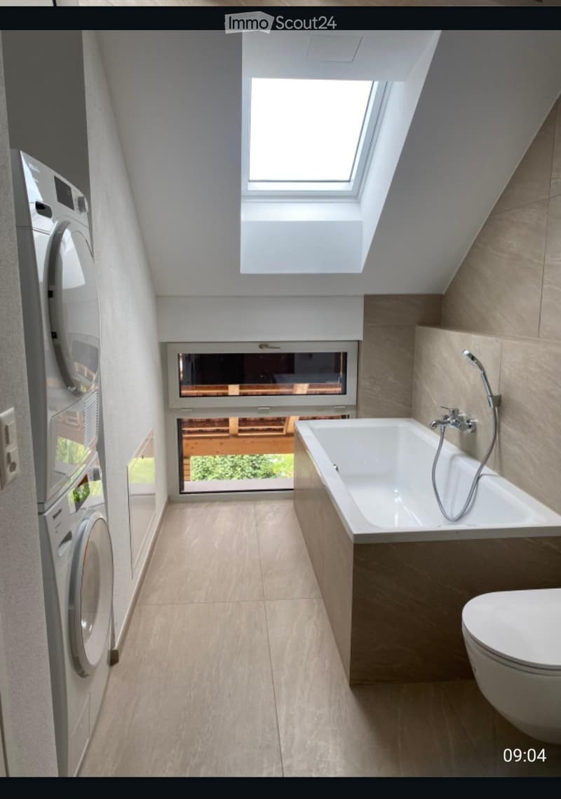 Modern bathroom with a bathtub, toilet, and washing machine. Features large windows and a skylight, with light-colored tiles and wooden accents.