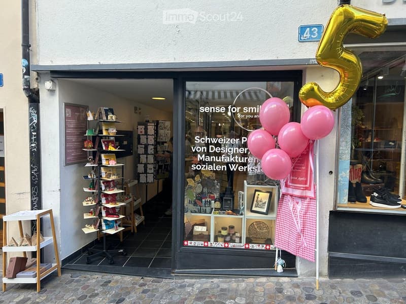 Storefront with glass windows displaying various items, including books, gifts, and shoes. A pink apron and balloons are visible outside the entrance.