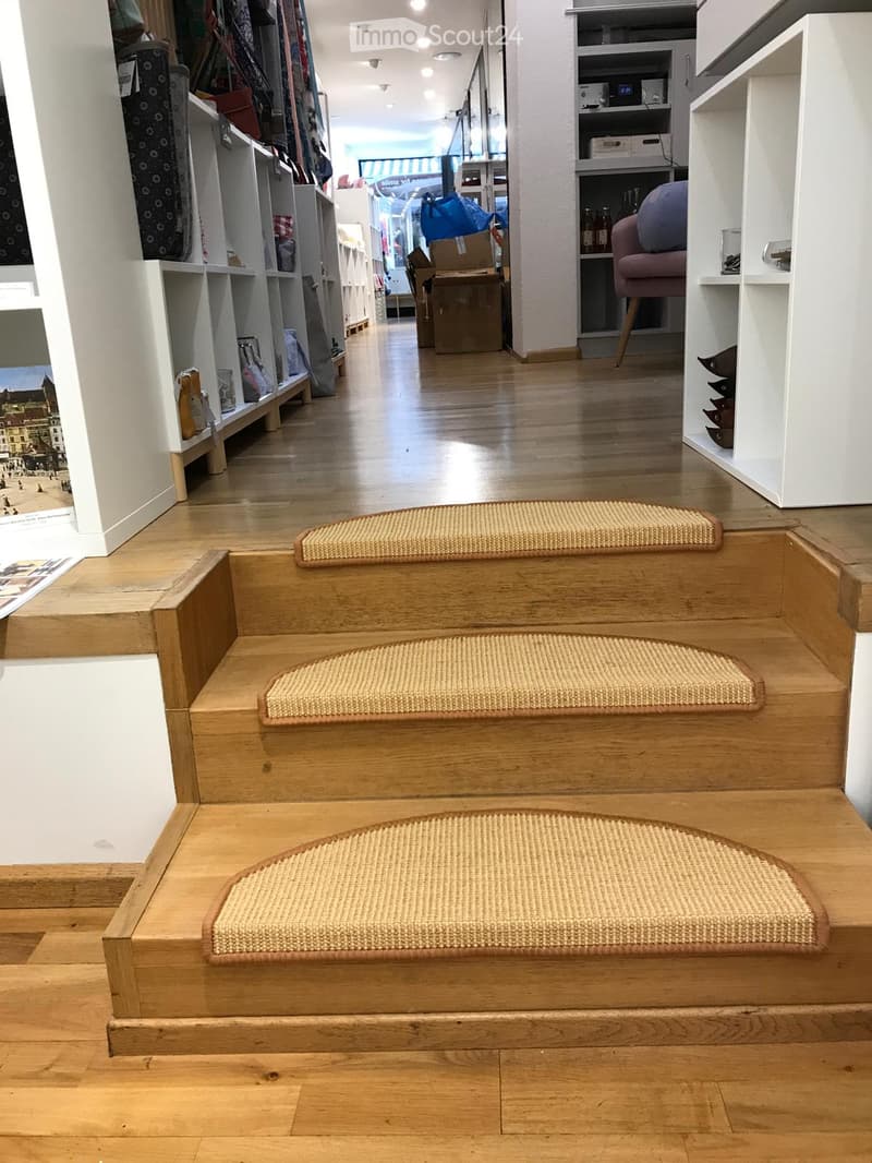 A well-lit hallway with wooden flooring, white walls, and built-in shelving units displaying various items such as clothing, bags, and decorative objects. There are also a few pieces of furniture, including a pink chair and a small table with bottles.