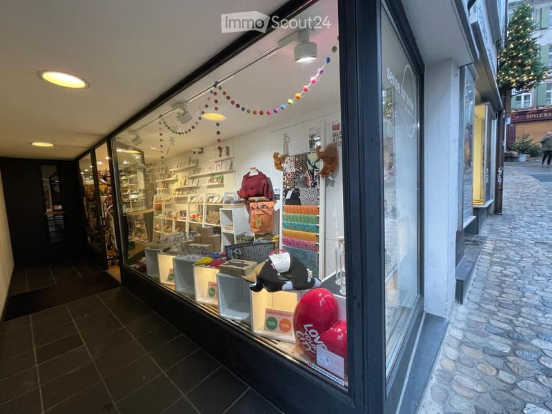 Shop interior visible through large glass windows, displaying shelves with books, a mannequin in clothing, handbags, balloons, and a sign offering discounts.