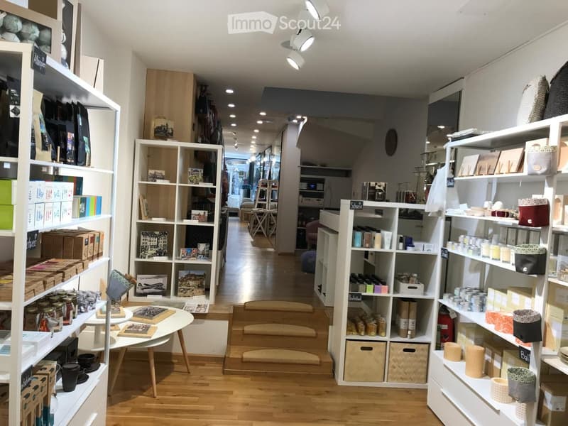 Well-lit store interior with wooden flooring, white shelving units filled with books, candles, jars, and boxes, a small round table with books and a bowl, and a few stools.