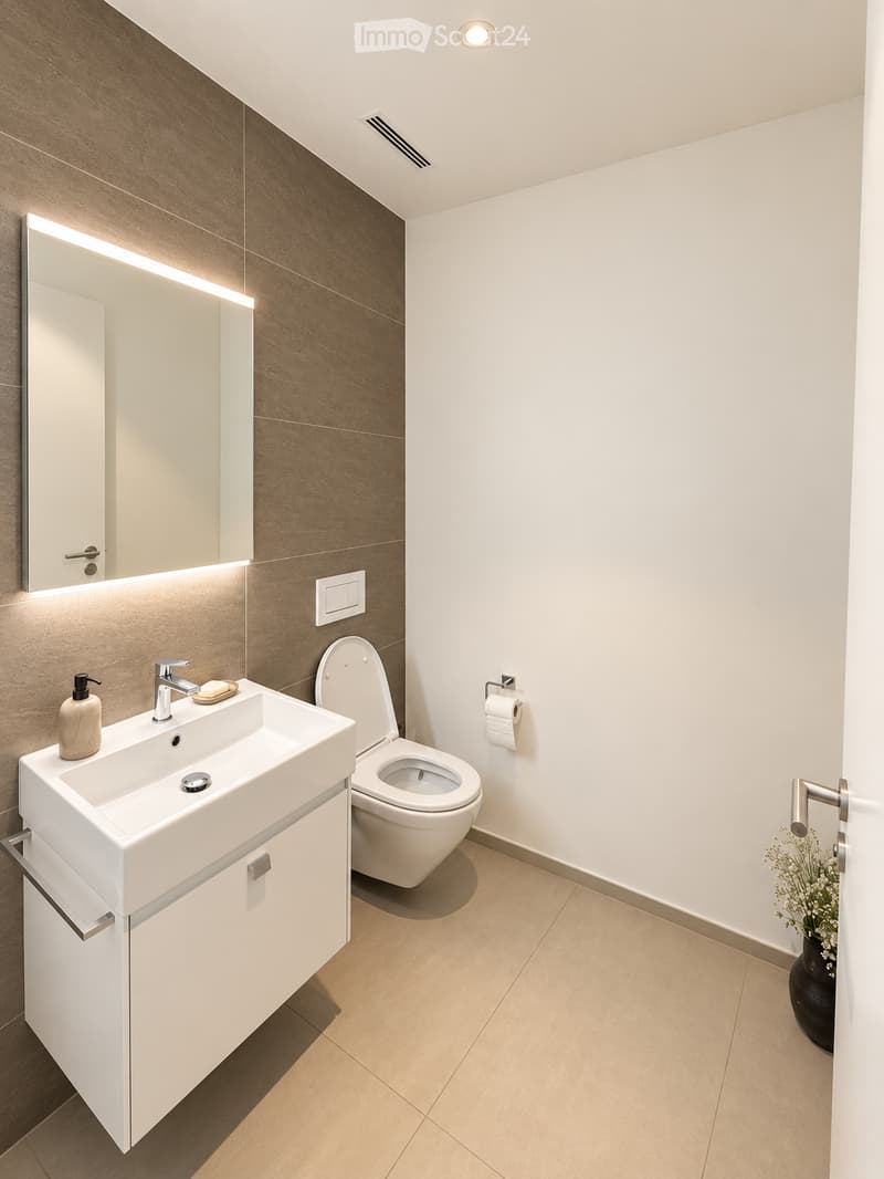 Modern bathroom with a white sink, toilet, large mirror with LED lighting, and a small vase with flowers on the floor.