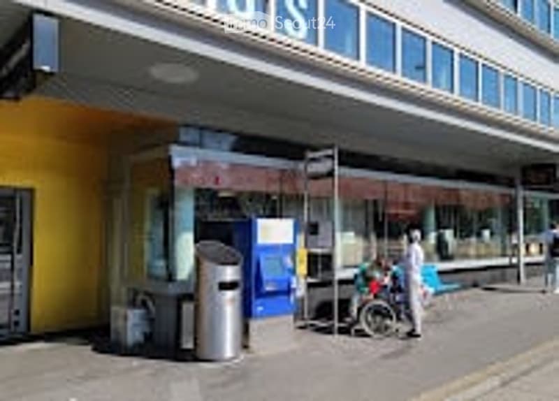 A modern building entrance with a yellow facade, glass windows, and a bus stop shelter nearby. There are a few people and a bicycle visible.