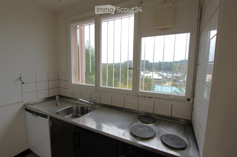 Empty kitchen with stainless steel sink, faucet, electric stove, and dishwasher, large windows overlooking a green area.