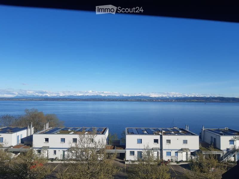 Modern white buildings with solar panels on the roof, situated near a large body of water with distant snow-capped mountains visible.