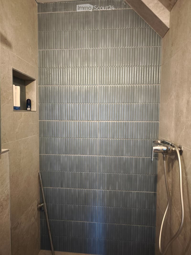 Shower area with grey tiled walls, a modern chrome faucet, and a small shelf holding toiletries.