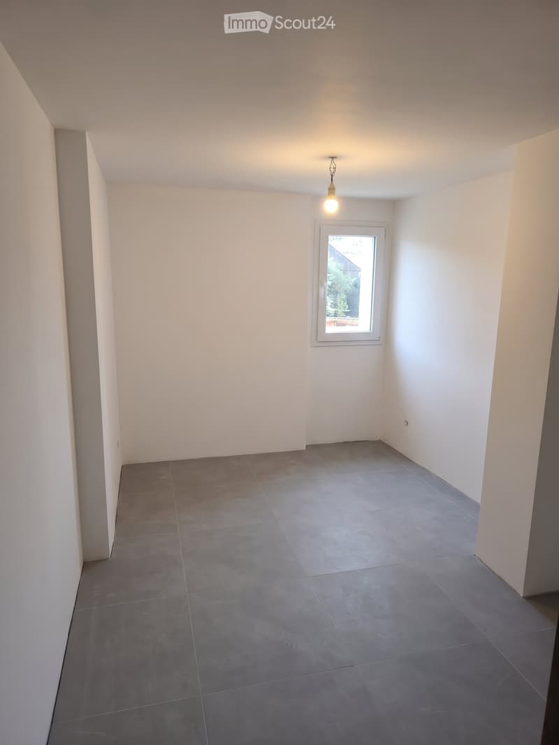 Empty room with grey tiled floor, white walls, and a single window with white frame. A single light fixture hangs from the ceiling.