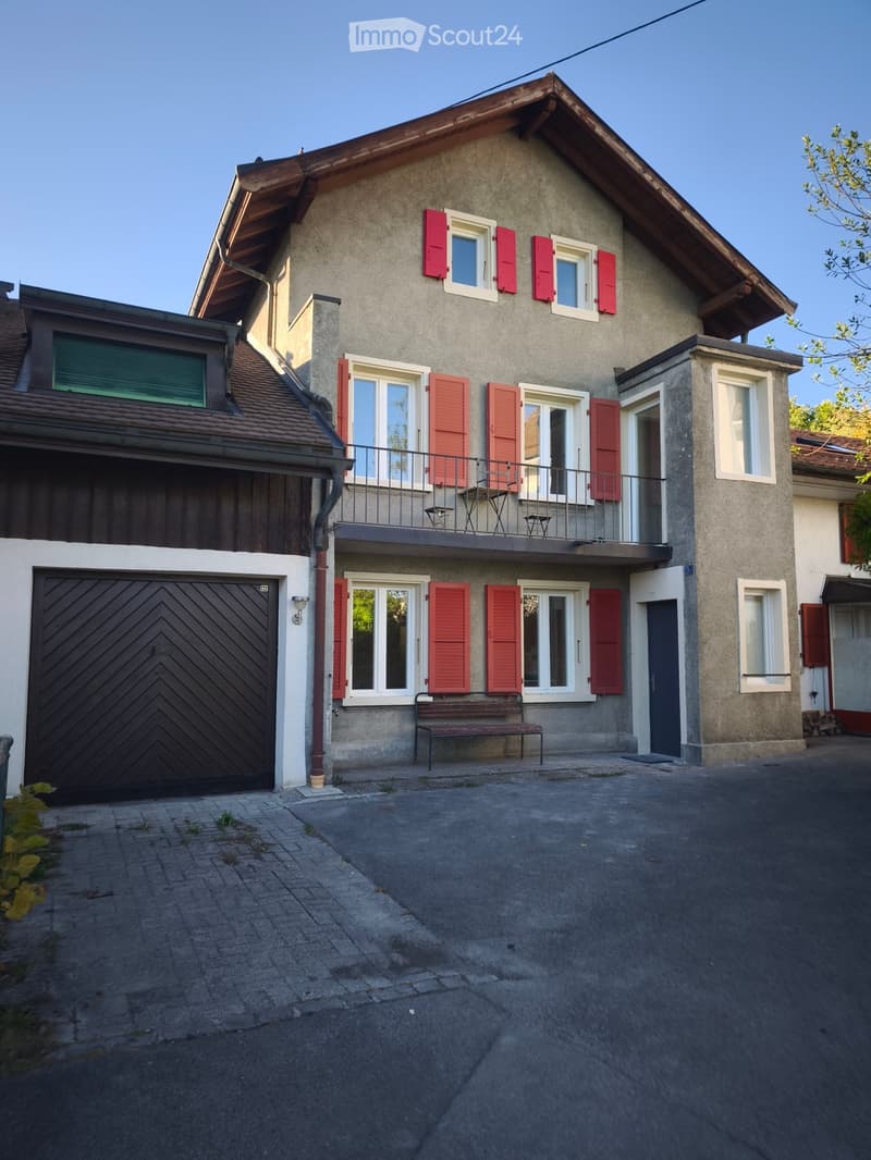 Two-story house with red shutters, white-framed windows, a balcony with outdoor furniture, and a garage. The house has a concrete facade and a slanted roof.