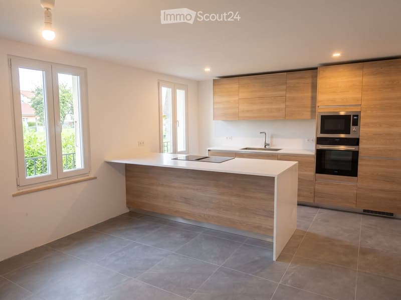 Modern kitchen with light wooden cabinets, white countertops, and stainless steel appliances including a microwave and oven. Features a central island with a built-in induction cooktop and ample natural light from large windows.