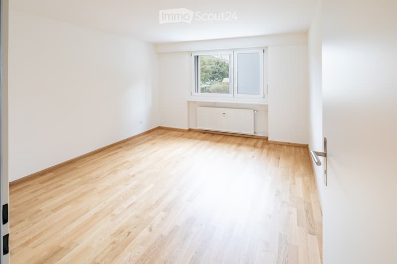 Empty room with light wooden flooring, white walls, and a window with a view of greenery outside. A radiator is visible below the window.