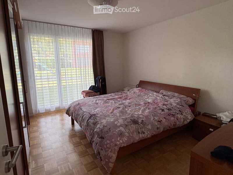 Bedroom with wooden flooring, large window with sheer curtains, bed with floral duvet, nightstand, and a small stool near the window.
