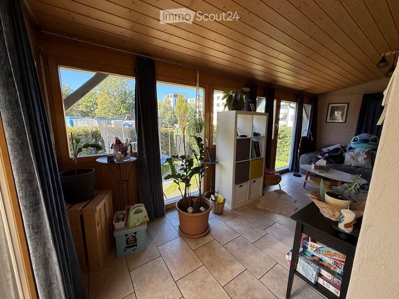 Bright, wooden-framed living room with large windows, a sofa, coffee table, bookshelf, and several potted plants.