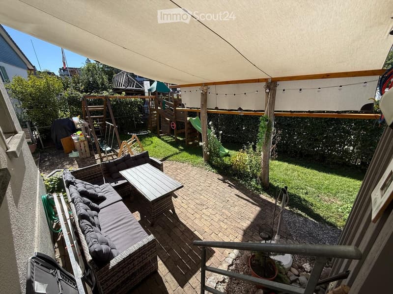 A covered outdoor patio with a wicker sofa, a wooden table, and a small grassy area with a slide and climbing frame.
