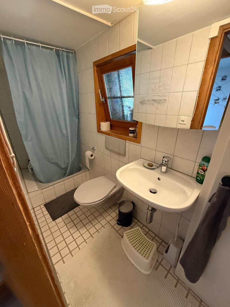 Compact bathroom with white tiles, a toilet, a sink with a mirror, a small window, and a shower area with a blue curtain.