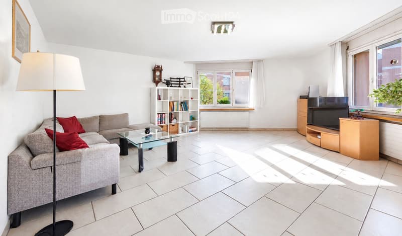 Bright living room with white walls, tiled floor, grey sofa with red cushions, glass coffee table, bookshelf, floor lamp, and TV on wooden stand.