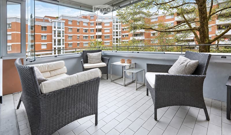 Balcony with wicker chairs, a small table, and cushions, overlooking a cityscape with modern buildings and trees.