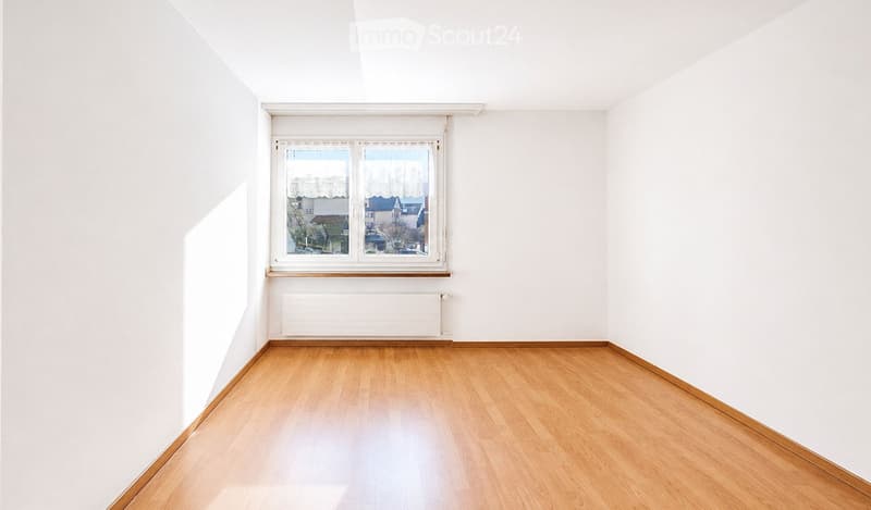 Empty room with white walls, wooden flooring, and a large window with a view of the outside. The room features a radiator below the window and a simple wooden baseboard along the walls.