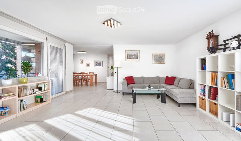 Bright living room with a grey sectional sofa, glass coffee table, bookshelf, dining area with chairs and table, and large windows letting in natural light.