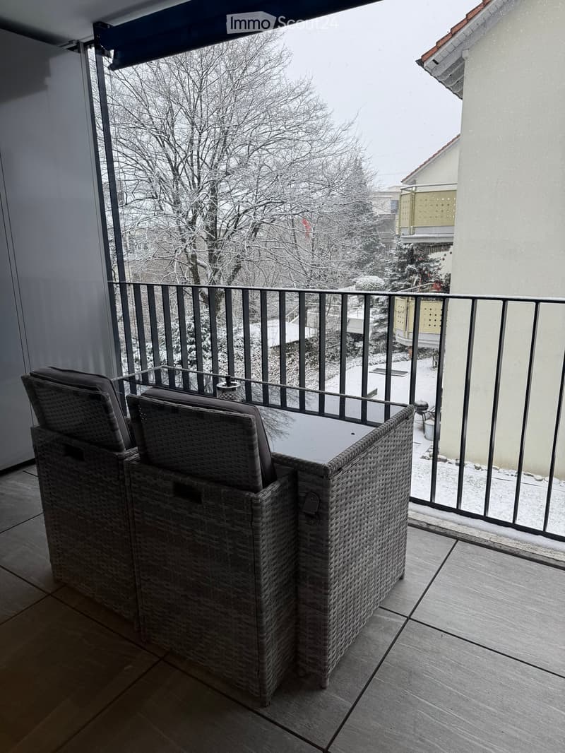 Balcony with two wicker chairs and a small table, overlooking a snowy landscape with trees and neighboring buildings.