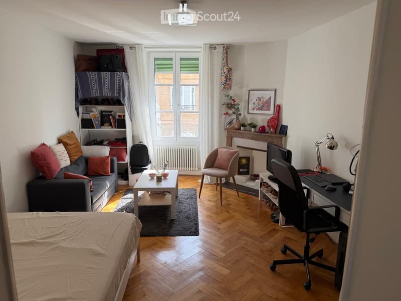 A multi-functional room with a bed, sofa, coffee table, desk, and chair, featuring wooden flooring and large windows with white curtains.