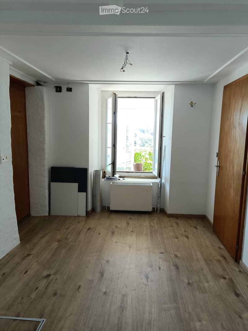 Empty room with wooden flooring, white walls, and a large window with open shutters. There is a radiator, a few stacked items near the window, and a wooden door on the right side.