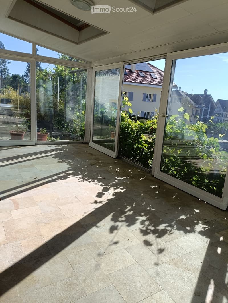 Bright, spacious balcony with large glass doors, tiled floor, and natural light. Shadows of plants are cast on the floor.