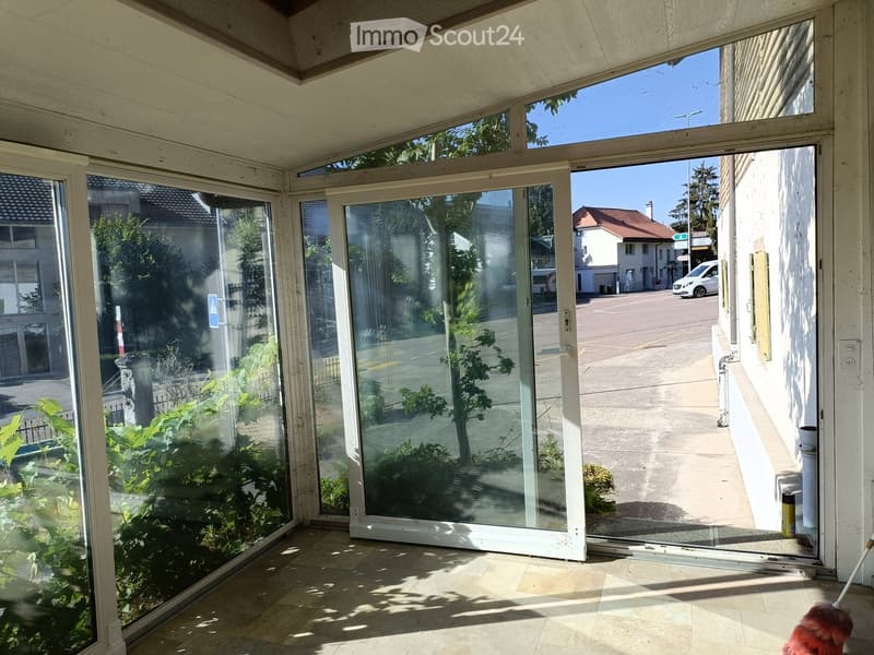 Bright, enclosed balcony area with large glass sliding doors, tiled floor, and visible outdoor greenery and street view.