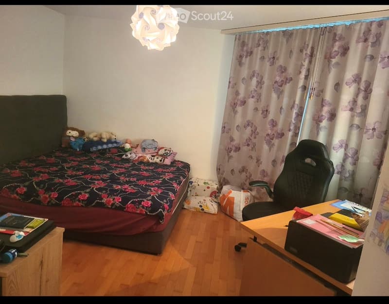 A cozy bedroom with a wooden floor, a bed with a colorful floral duvet, a desk with a chair, and a window with floral curtains. The room includes a lamp, books, and toys on the bed.