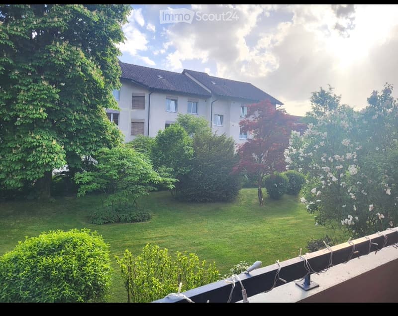 Balcony overlooking a well-maintained garden with a two-story house in the background, surrounded by trees and shrubs.
