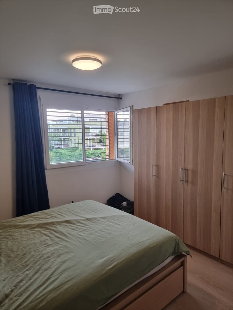 A modern bedroom with a wooden bed, a large wardrobe, and a window with blue curtains. A backpack is placed on the floor near the bed.
