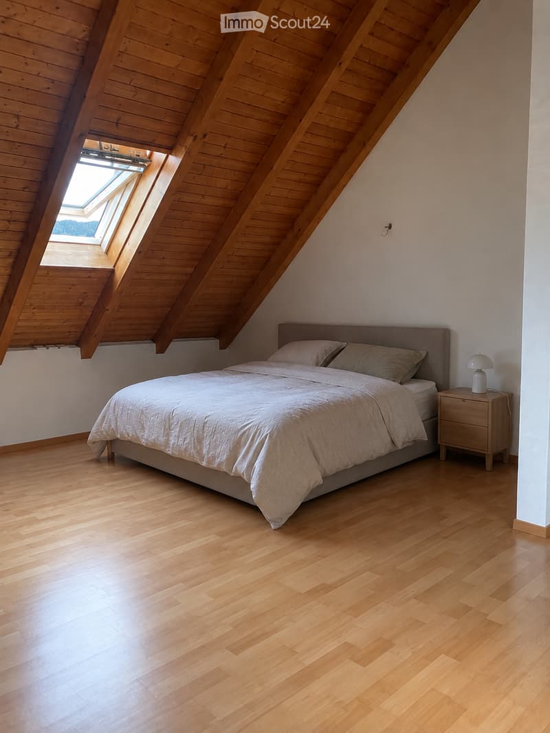 Bright, airy bedroom with wooden floors and slanted wooden ceiling. Features a large bed with white bedding, a nightstand with a lamp, and a skylight window bringing in natural light.